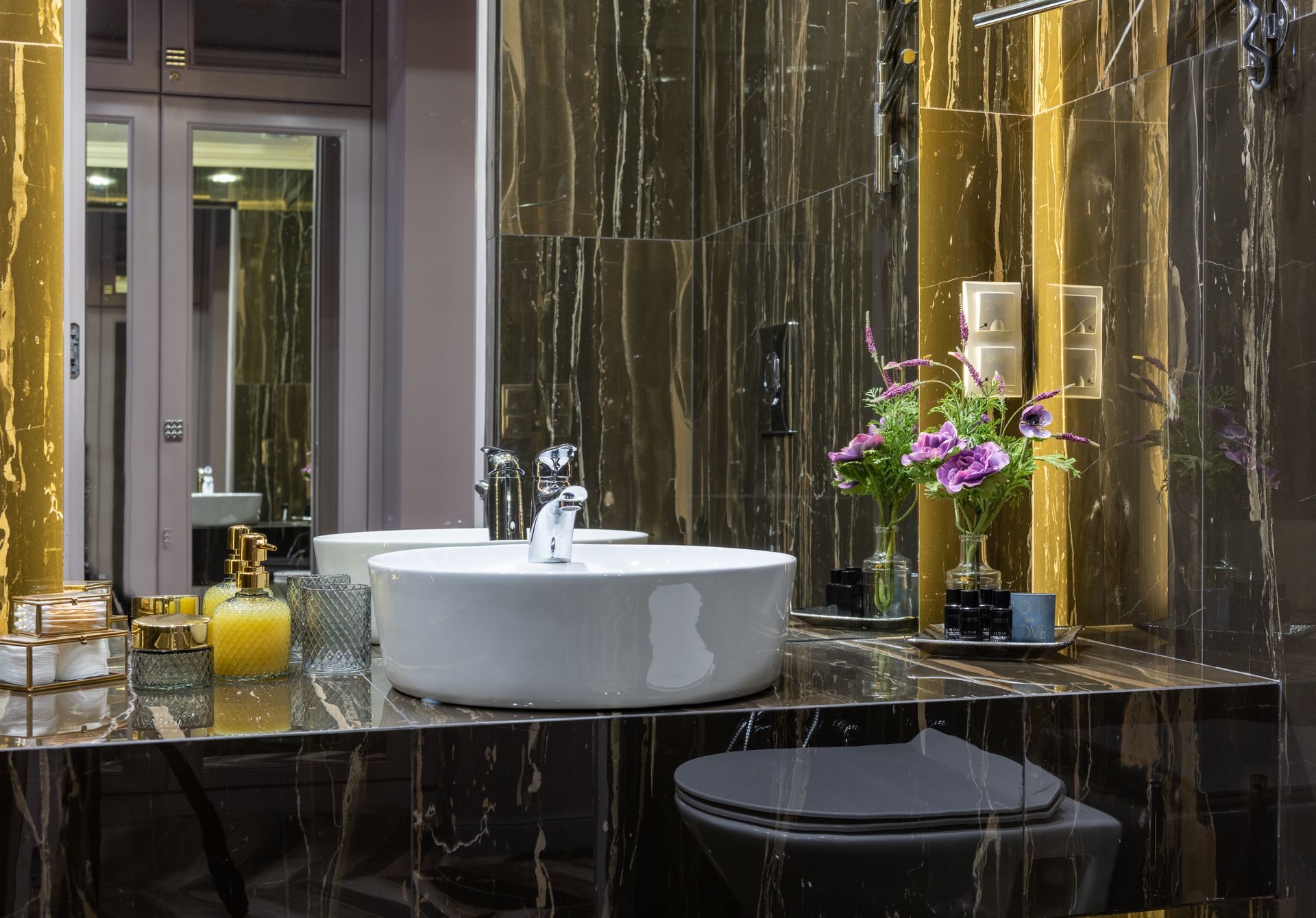 Contemporary bathroom with washstand between decorative flowers and liquid soap reflecting in mirror against door