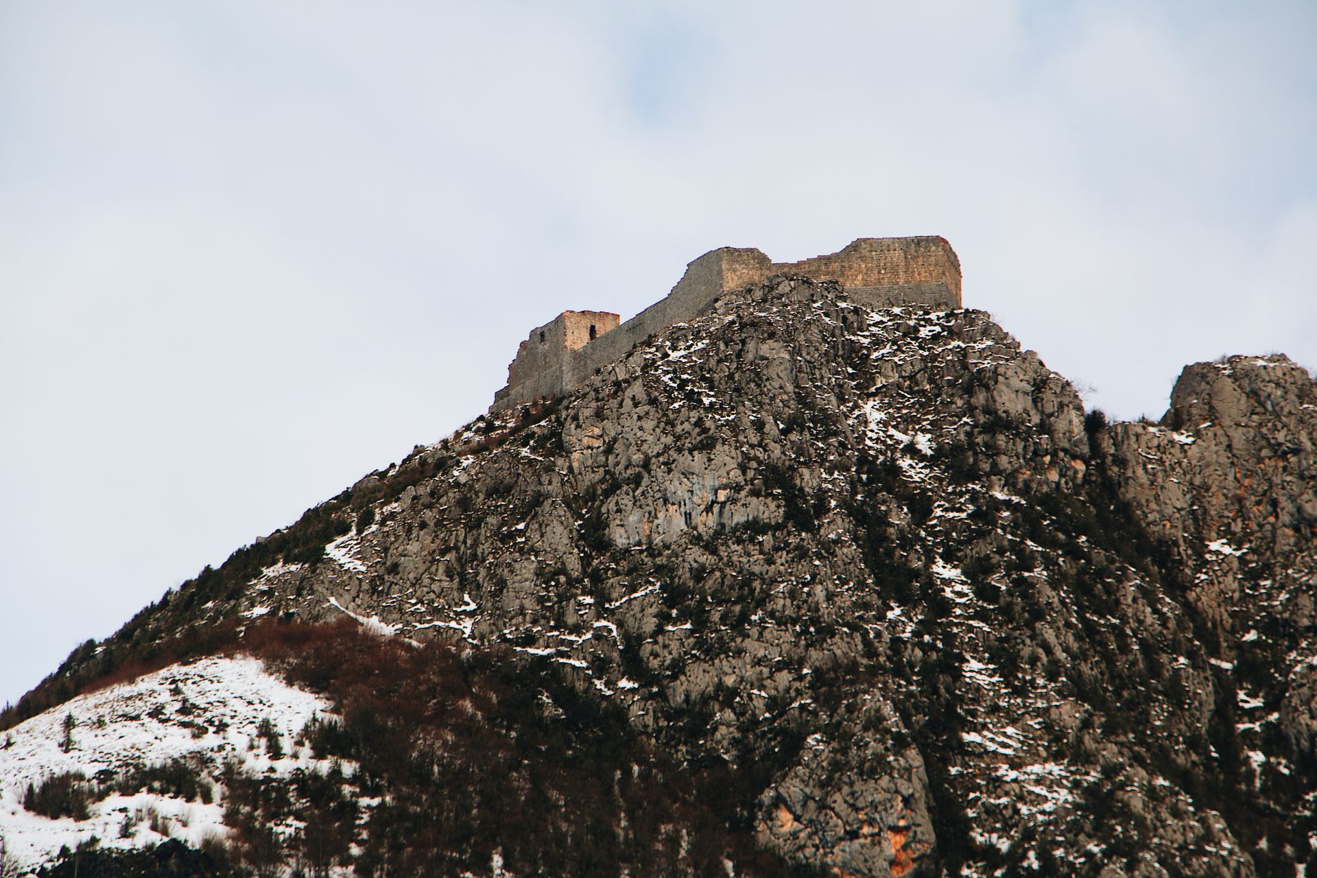A rugged, snow-dusted mountain with ancient ruins under a cloudy sky.