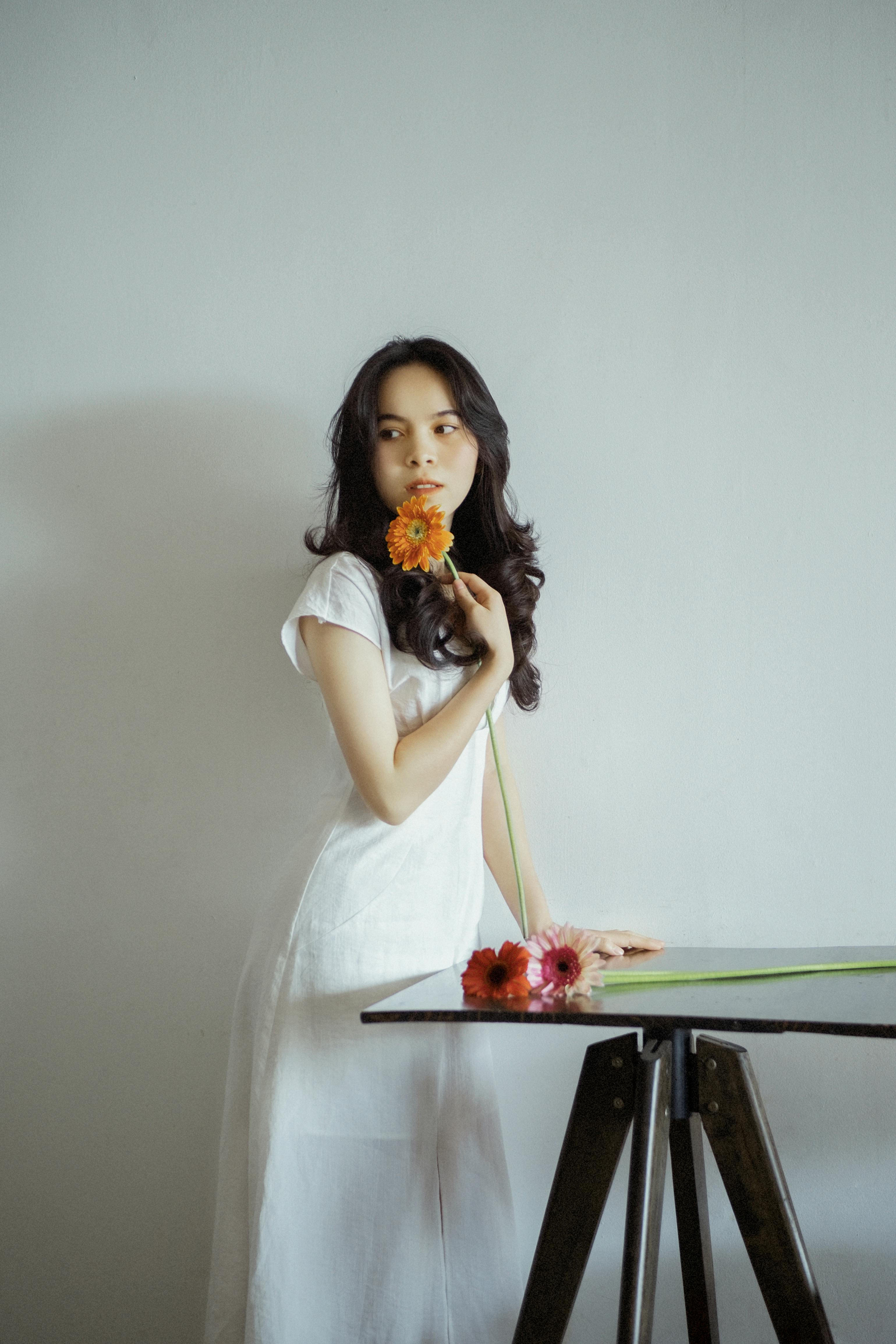 A young Asian woman in a white dress poses with flowers in a minimalist setting, capturing a serene and elegant moment.