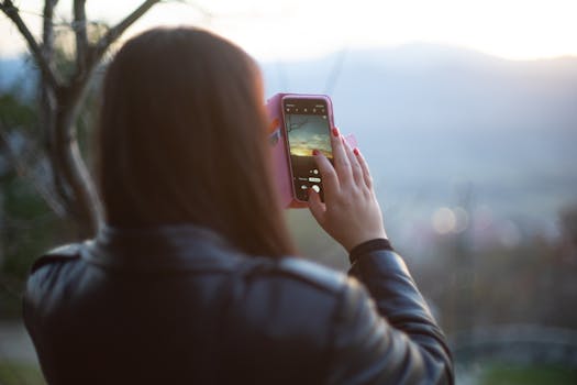 A woman in a leather jacket captures a scenic sunset view using her smartphone outdoors.