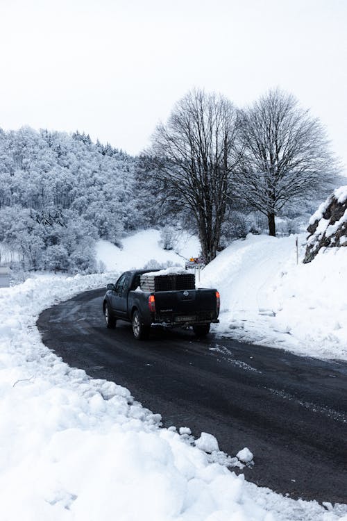 Modern pickup truck driving on curvy asphalt road amidst snowy hills covered with lush trees on cloudy winter day