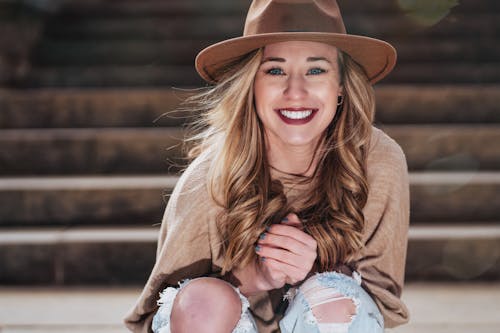 Happy attractive female in casual outfit and hat sitting with hands clasped on outdoor stairway and looking at camera with toothy smile