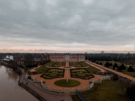 Drone view of historic Hampton Court Palace and gardens on a cloudy day, showcasing its grandeur and serene beauty. by Ollie Craig