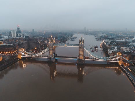 Aerial view of London city located in England with illuminated Tower Bridge on River Thames near modern buildings under gray cloudy sky in foggy day