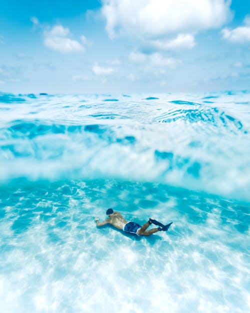 Full body of anonymous male diver with slippers swimming under transparent blue water with sun glares in tropical lagoon against cloudy sky
