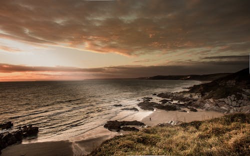 Rocky sandy coast with green grass washed by waves of endless water of ocean under vast cloudy sky