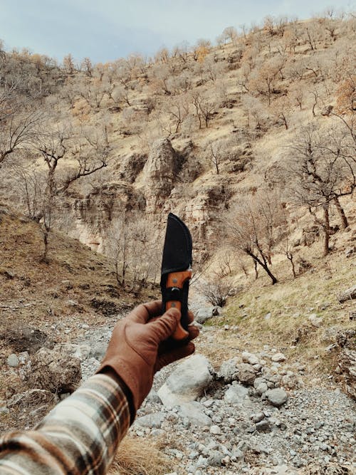 Crop anonymous hiker wearing leather glove demonstrating sharp knife in hand while standing near hilly area with trees in nature