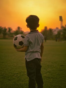 A young boy stands with a soccer ball in a field during a warm sunset.