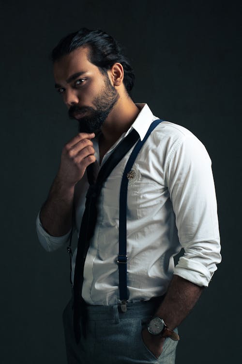 Serious ethnic bearded young male in white shirt with tie and pants with suspenders and wristwatch standing on gray background and looking at camera with hand at chin