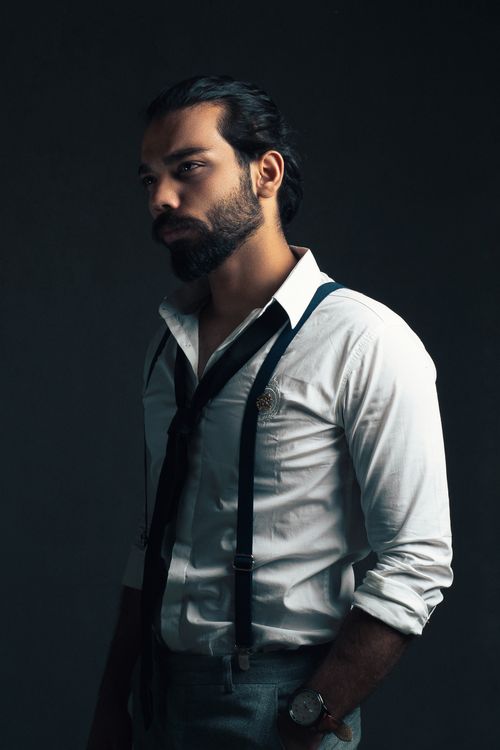 Free Confident bearded ethnic young businessman in white shirt with tie and pants with suspenders and wristwatch standing in studio on gray background and looking away with hand in pocket Stock Photo