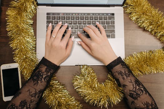 Close-up of hands typing on a laptop with festive gold tinsel decoration, creating a holiday work atmosphere.