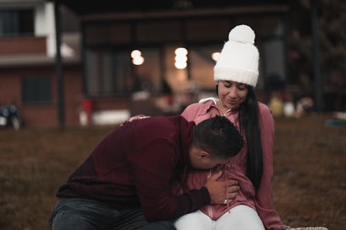 Free Content male in warm sweater tenderly kissing content wife belly wearing warm hat while sitting together on grassy meadow on backyard Stock Photo