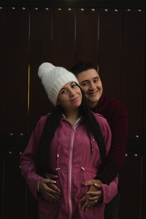 Free Content ethnic man standing behind and hugging young pregnant wife in warm wear while standing together near fence and looking at each other Stock Photo