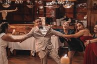 Bride in wedding dress pulling shocked groom away from positive girlfriends while having fun during wedding celebration