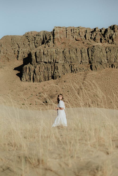 Dreamy female in white dress standing on sandy ground against high rocky formations under cloudless blue sky in sunny day