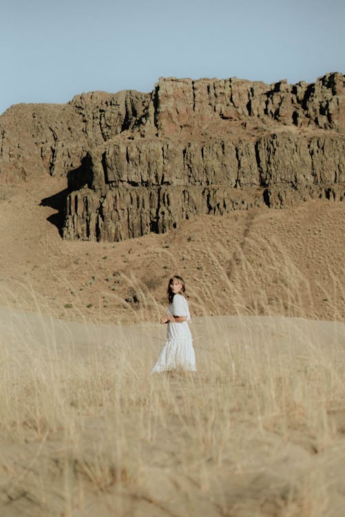Free Dreamy female in white dress standing on sandy ground against high rocky formations under cloudless blue sky in sunny day Stock Photo