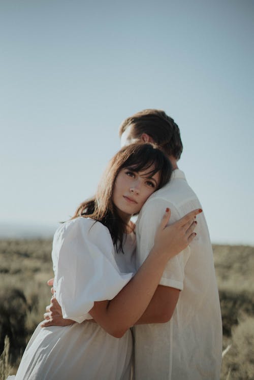 Calm female in light dress embracing and leaning on shoulder of boyfriend while standing in field in countryside in sunny day