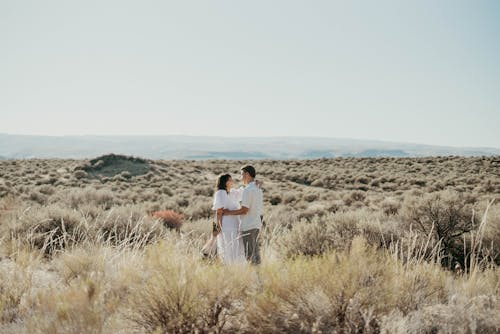 Cheerful couple in stylish clothes hugging while standing in field with dry bushes in countryside and looking at each other