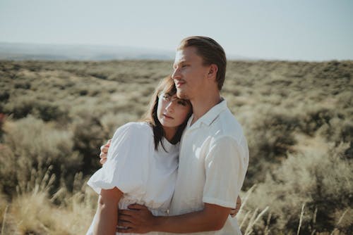 Cheerful couple in stylish light clothes hugging in field with dry grass in countryside and looking away in sunny day