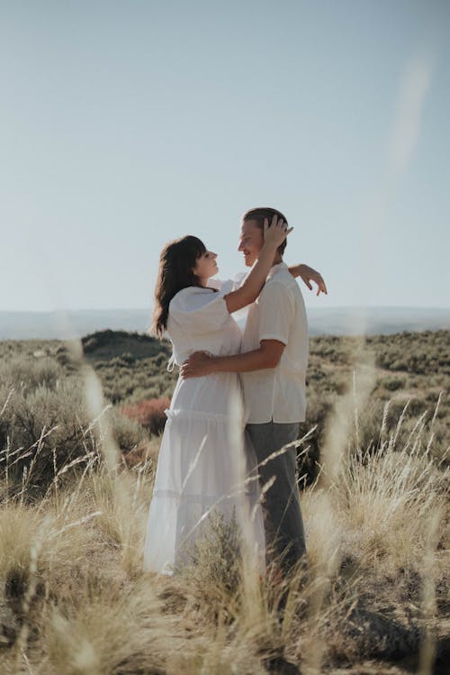 Side view of happy couple in stylish clothes embracing and looking at each other while standing in field with dry grass under cloudless blue sky in daytime