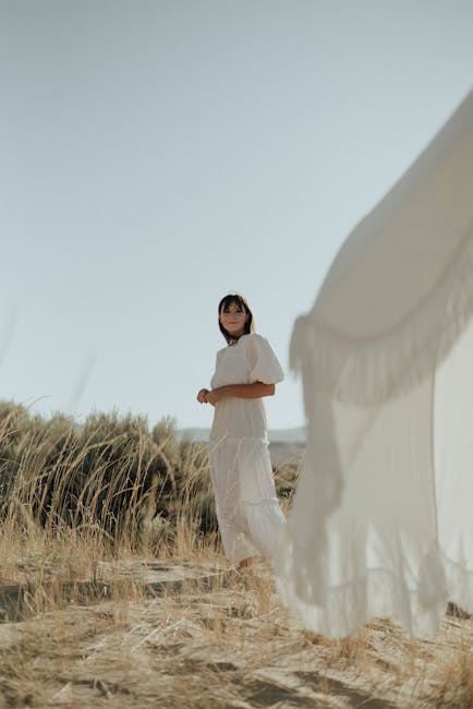Low angle of young woman in elegant white dress standing near white tent in rural grassy field