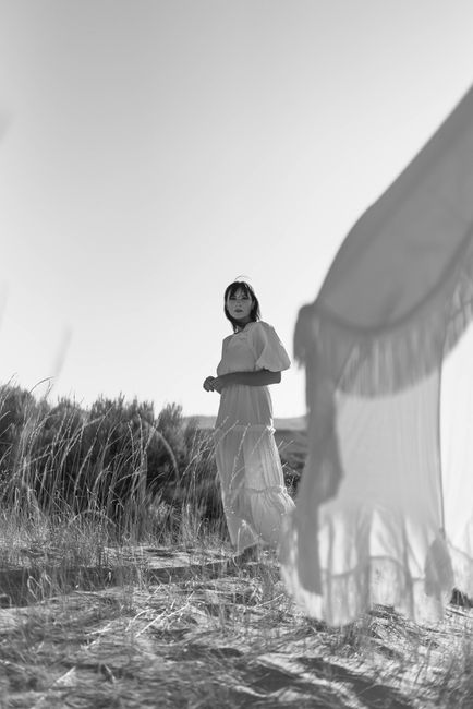 Full body of young woman in long white dress standing beside white tent in rural field in summer