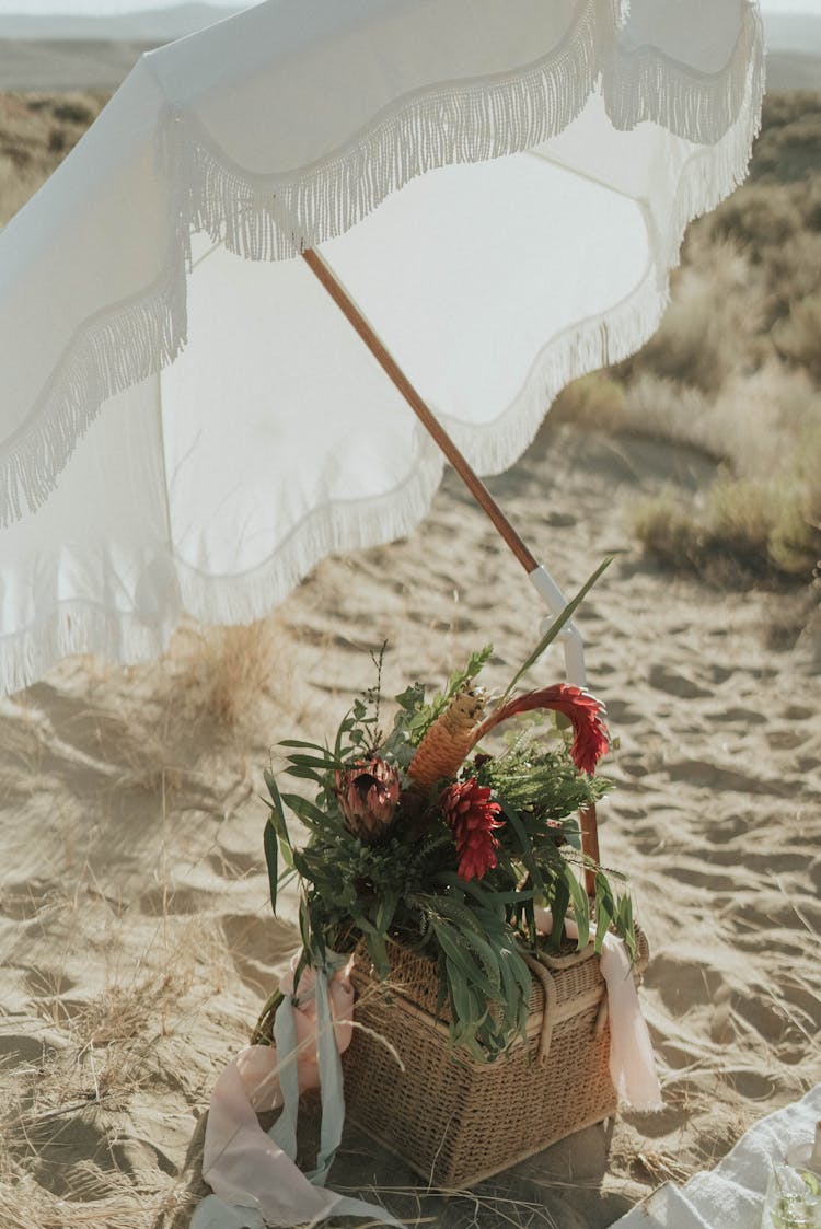 Basket With Flowers And Umbrella On Sandy Beach