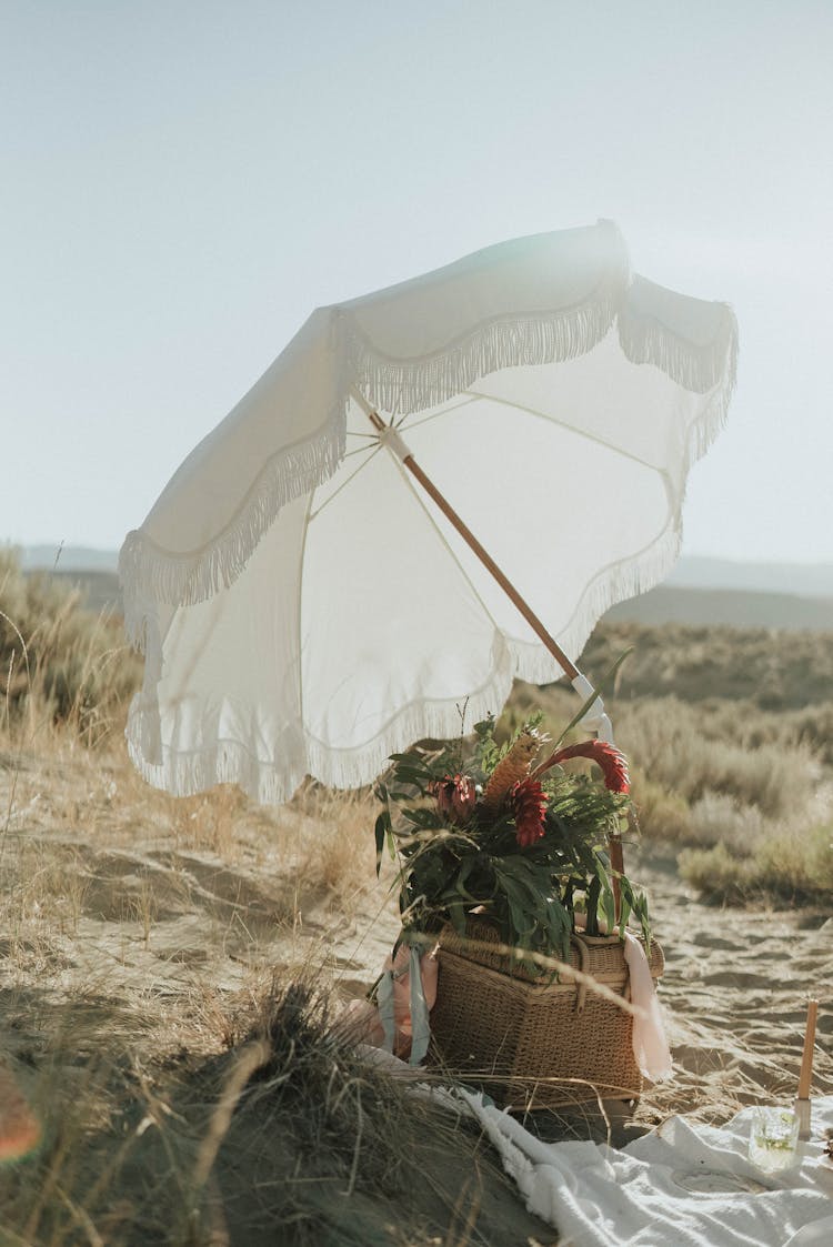 White Umbrella And Basket With Flowers On Sandy Coast