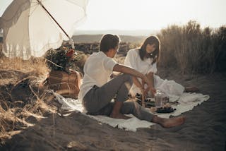 Young loving couple having romantic picnic sitting on white blanket with food and drinks under white umbrella