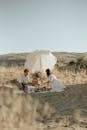 Young couple in white clothes sitting together on sandy dune and having picnic under white umbrella