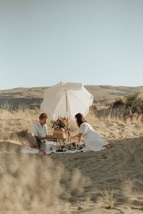 Free Young couple in white clothes sitting together on sandy dune and having picnic under white umbrella Stock Photo