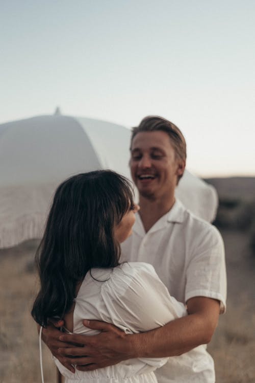 Happy couple in white clothes standing in dry desert and embracing gently under bright sky in daytime near umbrella