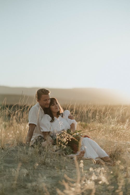 Young couple in white clothes sitting on dry grass in sandy field and embracing gently in sunny summer day under bright cloudless sky in countryside