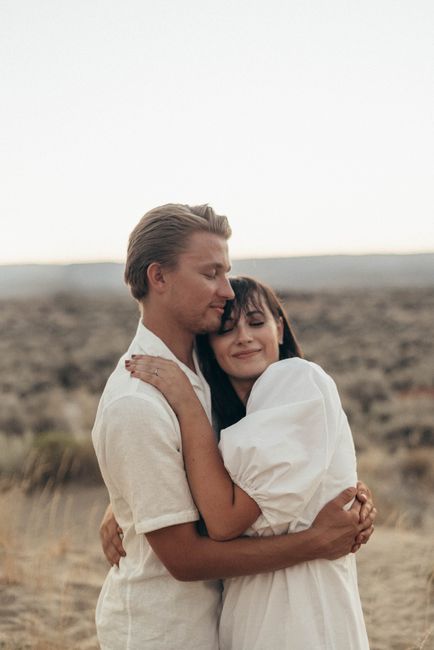Side view of positive young couple in white clothes standing near dry grass in sandy meadow and hugging in daylight under bright sky in nature