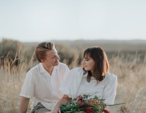 Positive young couple in white clothes with bouquet of flowers in hands sitting in field on dry grass under bright cloudless sky in sunny summer day with closed eyes