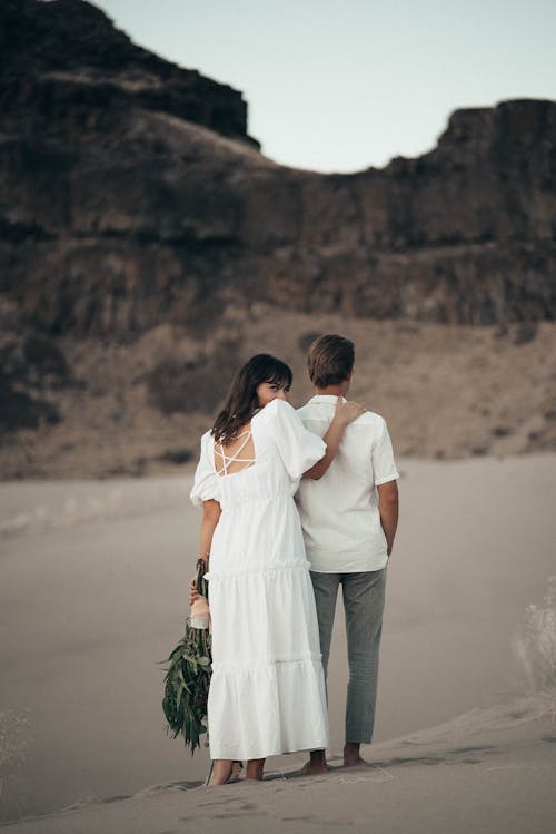 Full body of loving bride with bouquet of  flowers looking at camera over shoulder while touching shoulder of anonymous groom during wedding celebration