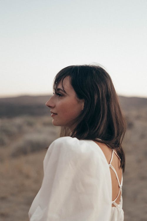 Side view of dreamy female in elegant white dress looking into distance while standing on grassy meadow on blurred background