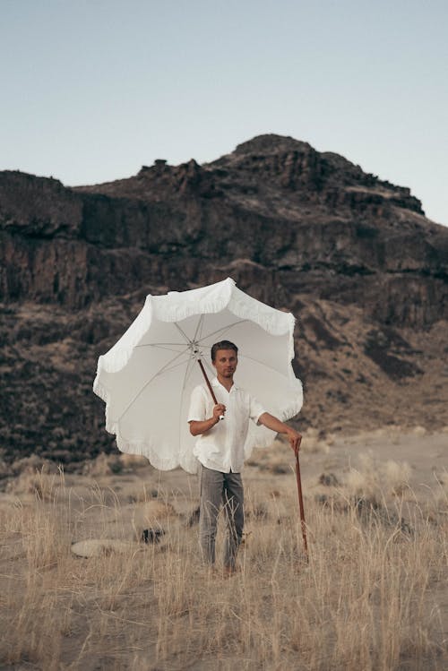 Full body of elegant male with white parasol and cane standing in meadow with dry grass against rocky mountain in nature