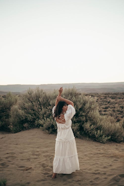 Full body back view of barefoot anonymous female in elegant white dress standing on dry ground near bushes while dancing with raised arms