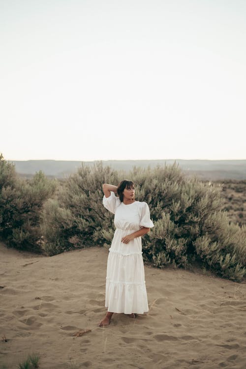 Full body of barefoot female wearing elegant white dress standing with hand behind head on dry ground near green bushes in countryside