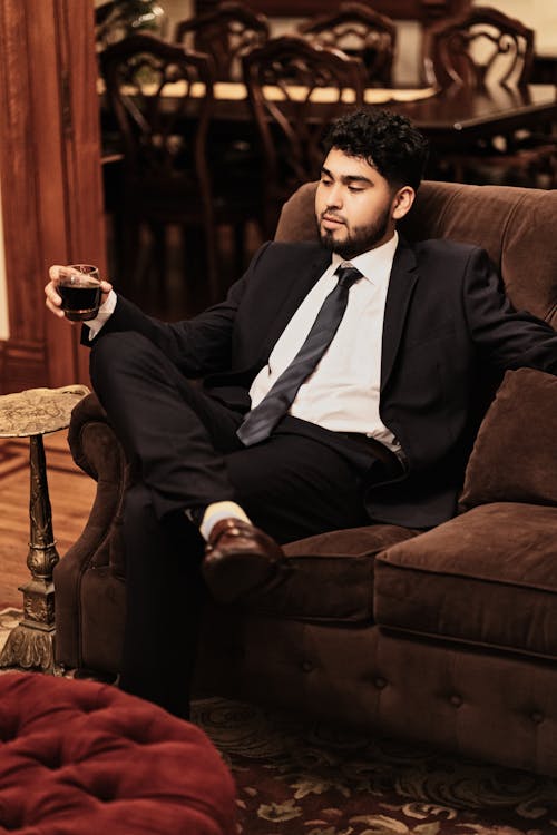 Confident well dressed ethnic male in classy suit sitting on cozy couch and enjoying aromatic freshly brewed coffee