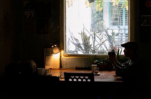 Side view of anonymous male sitting at wooden table with lamp near window in dark room in daytime