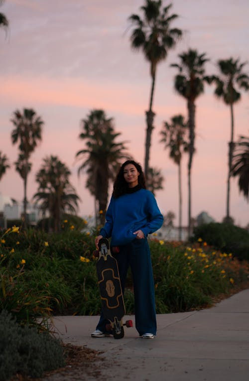 Full body of young female in casual clothes standing with skateboard on sidewalk near green grass and palms and looking at camera in summer evening
