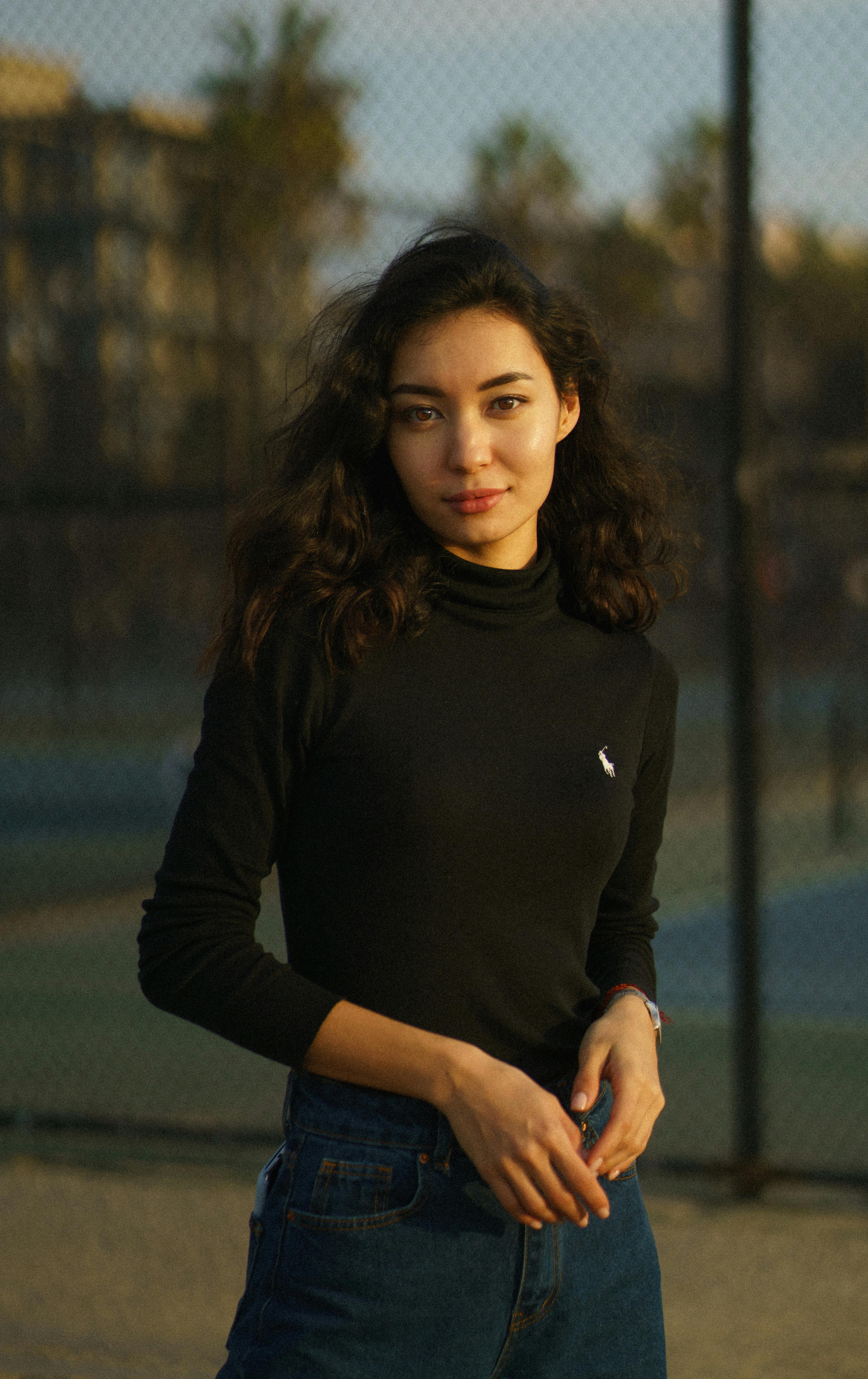 Young contemplative calm female in jeans and black blouse standing against metal net fence and looking at camera
