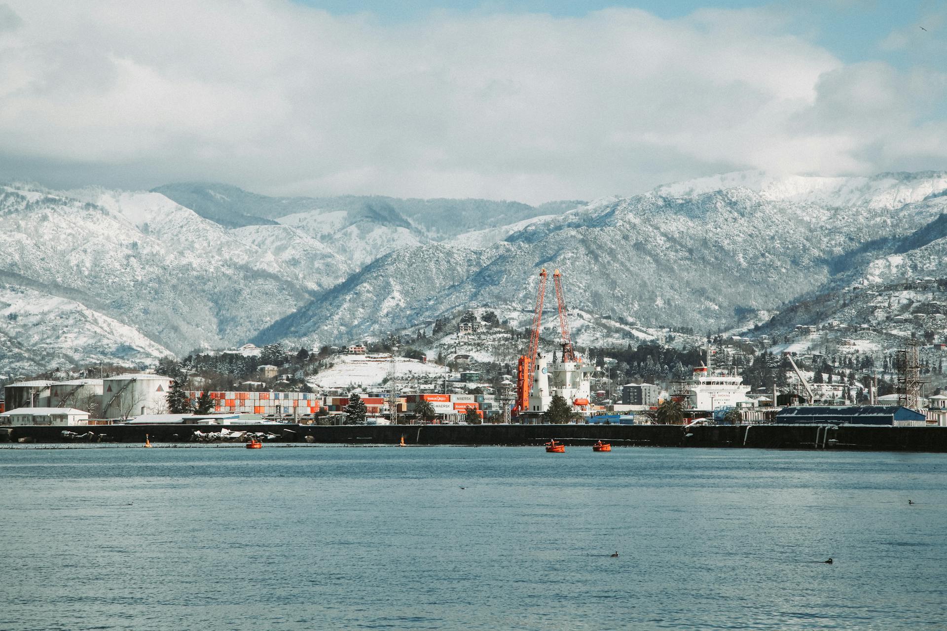 Port with cranes and industrial constructions located on shore near rippling water and hilly terrain covered with snow in winter time