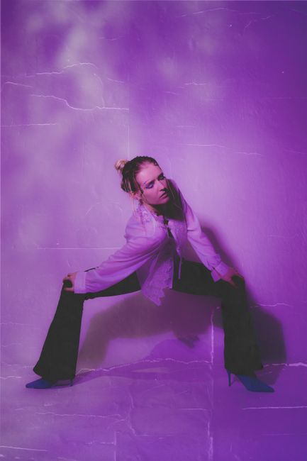 Full body of thoughtful young female in stylish outfit standing in illuminated purple room leaning on knees with closed eyes