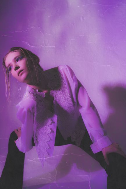 Gorgeous young lady in stylish outfit standing legs apart and leaning on knees in room with bright purple neon illumination