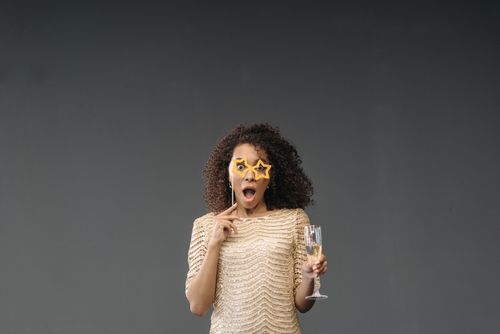Free Astonished woman in gold outfit with star-shaped glasses, holding champagne glass against a plain background. Stock Photo