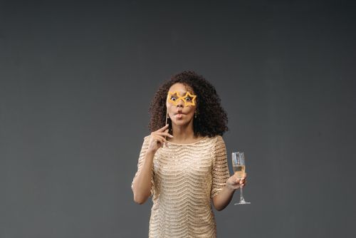 Free A cheerful woman in a sparkling dress poses with star-shaped glasses and a glass of champagne in a studio setting. Stock Photo