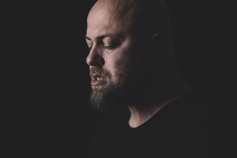 Close-up moody portrait of a bearded bald man in a dark studio setting.